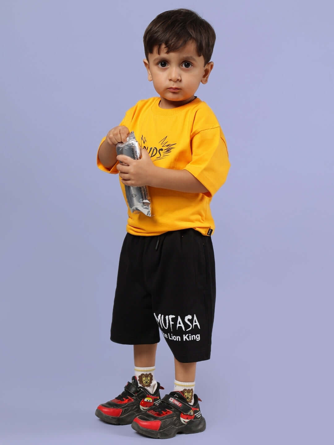 Young child wearing a mustard MUFASA T-shirt and black shorts, styled for boys and girls, standing against a gray background.
