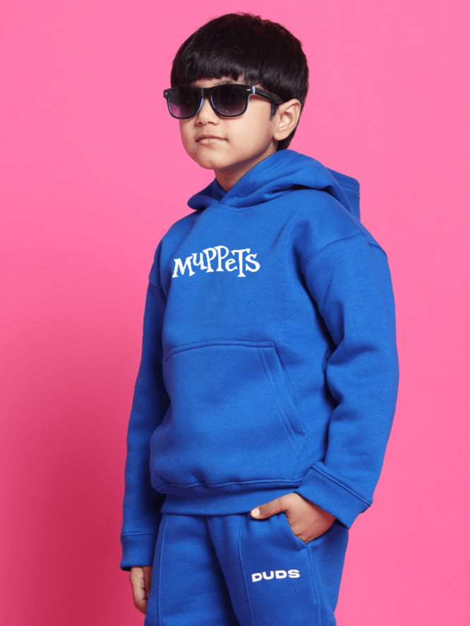 Child wearing a blue Muppets oversized hoodie and sunglasses against a pink background.