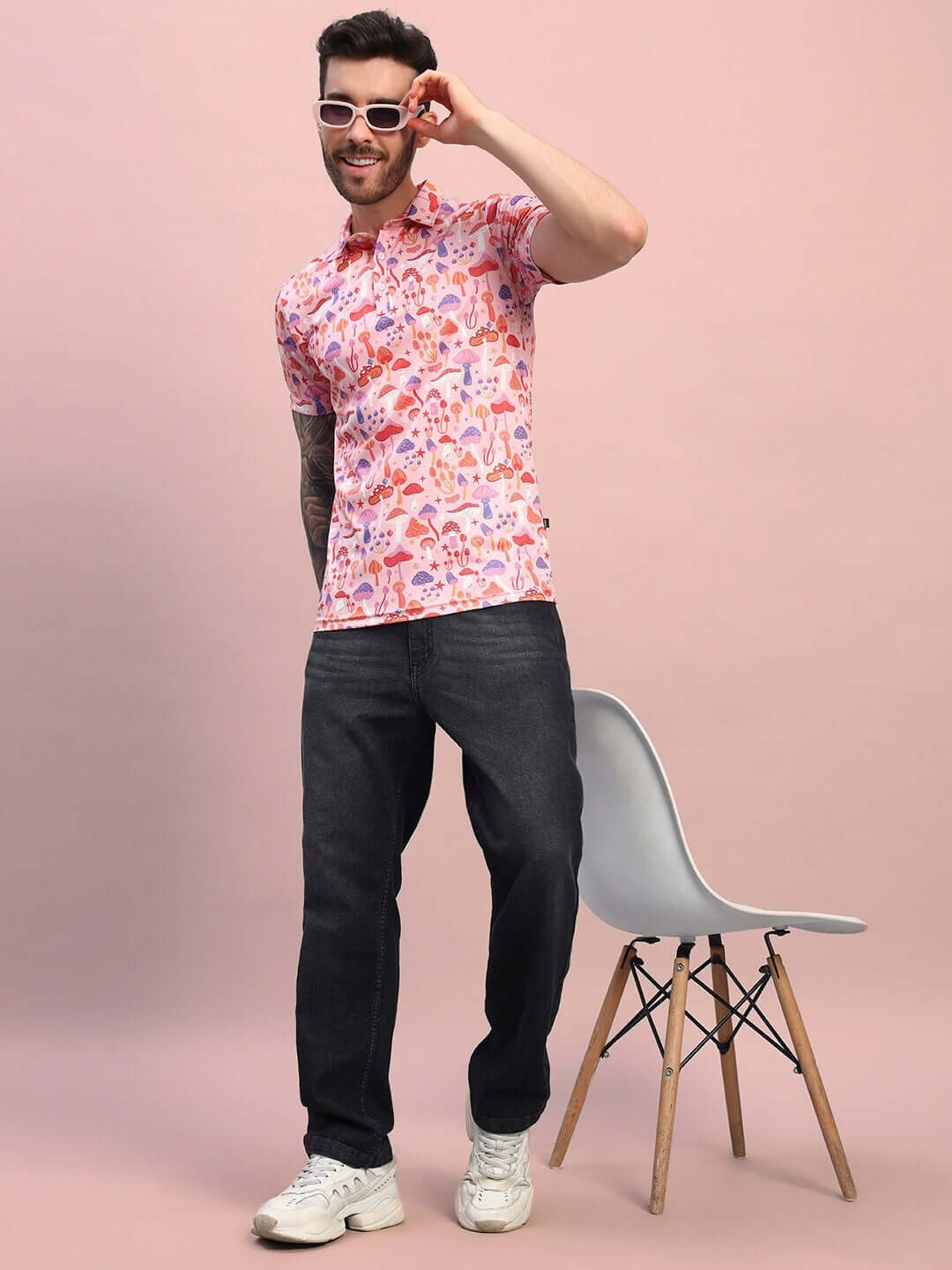 Man wearing a mushroom print regular fit t-shirt in pink multi-color, styled with dark jeans and white sneakers, standing beside a chair.