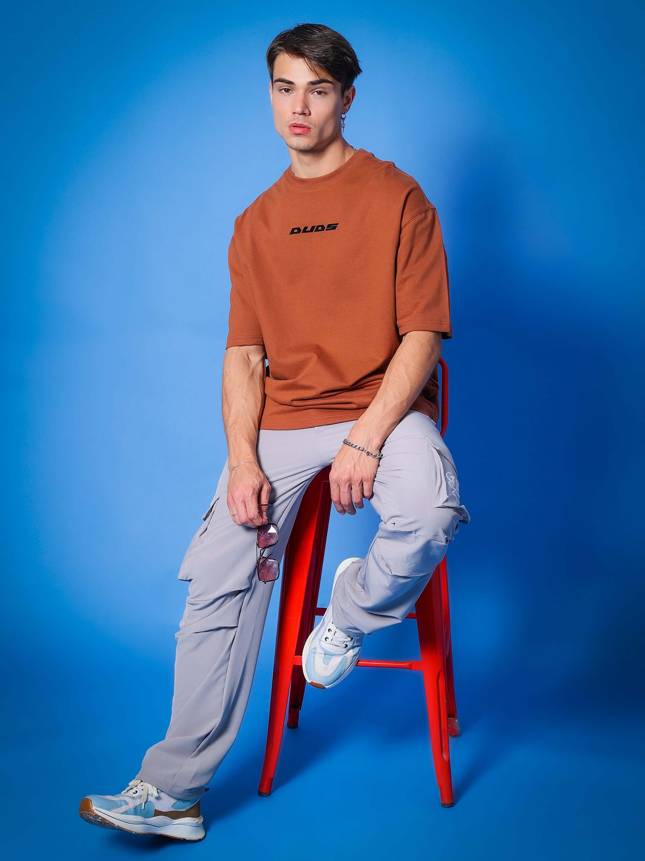Model wearing MYRIAD Relaxed Fit Cargo Pants in grey with a brown tee, seated on a red stool with a blue background.
