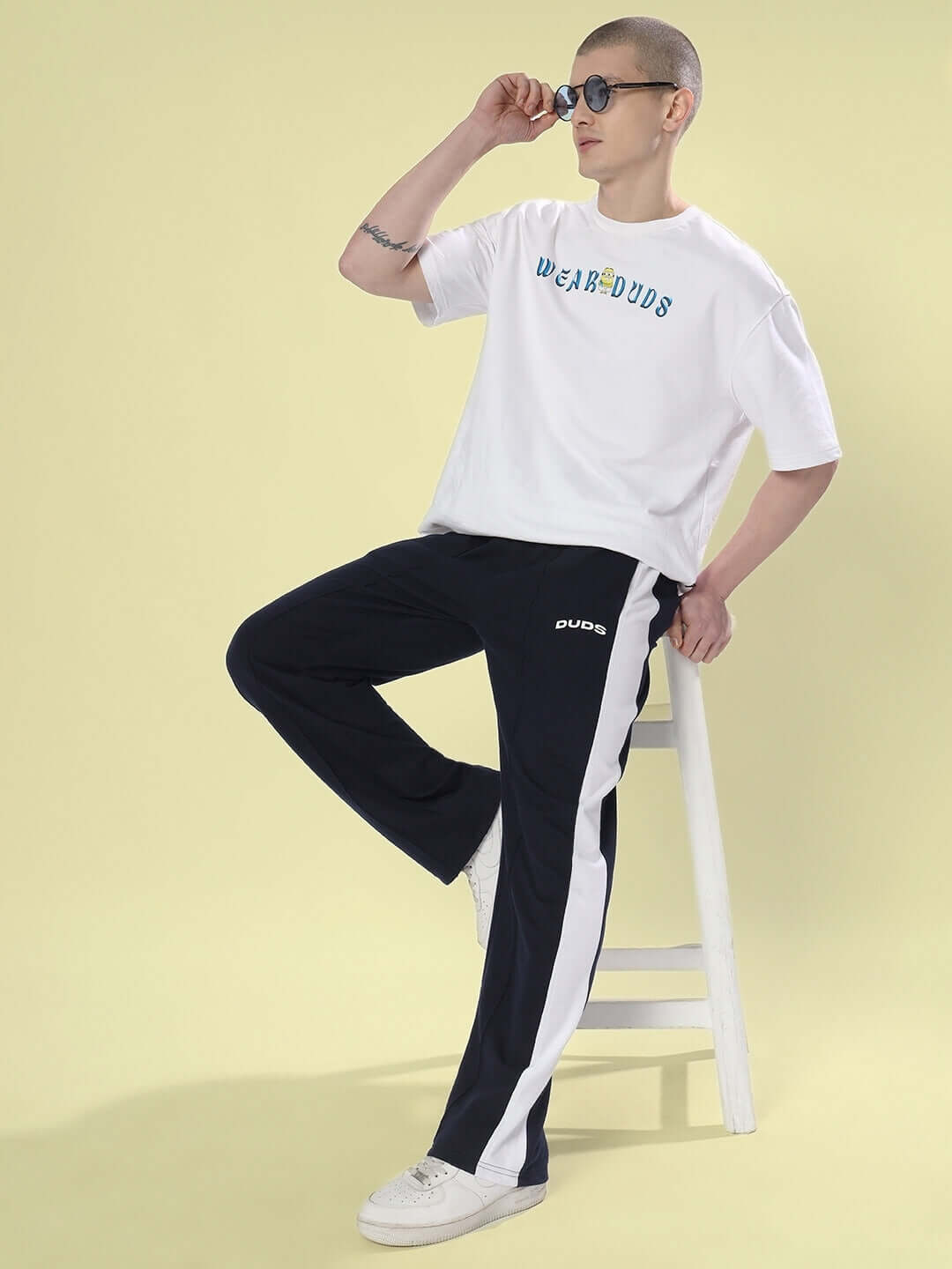 Model wearing navy blue side seam plated joggers by DUDS, sitting on a stool, paired with a white T-shirt and sunglasses.