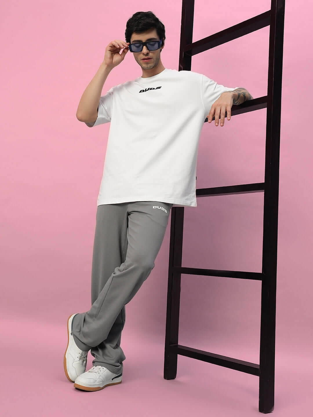 Model wearing New Era Relaxed Fit Grey Joggers by DUDS, styled with a white t-shirt and sunglasses, posing against a pink background.