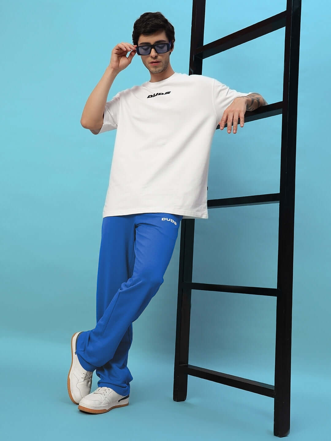 Man wearing the NEW ERA RELAXED FIT JOGGERS ROYAL BLUE paired with a white t-shirt, posing next to a ladder in a stylish setting.