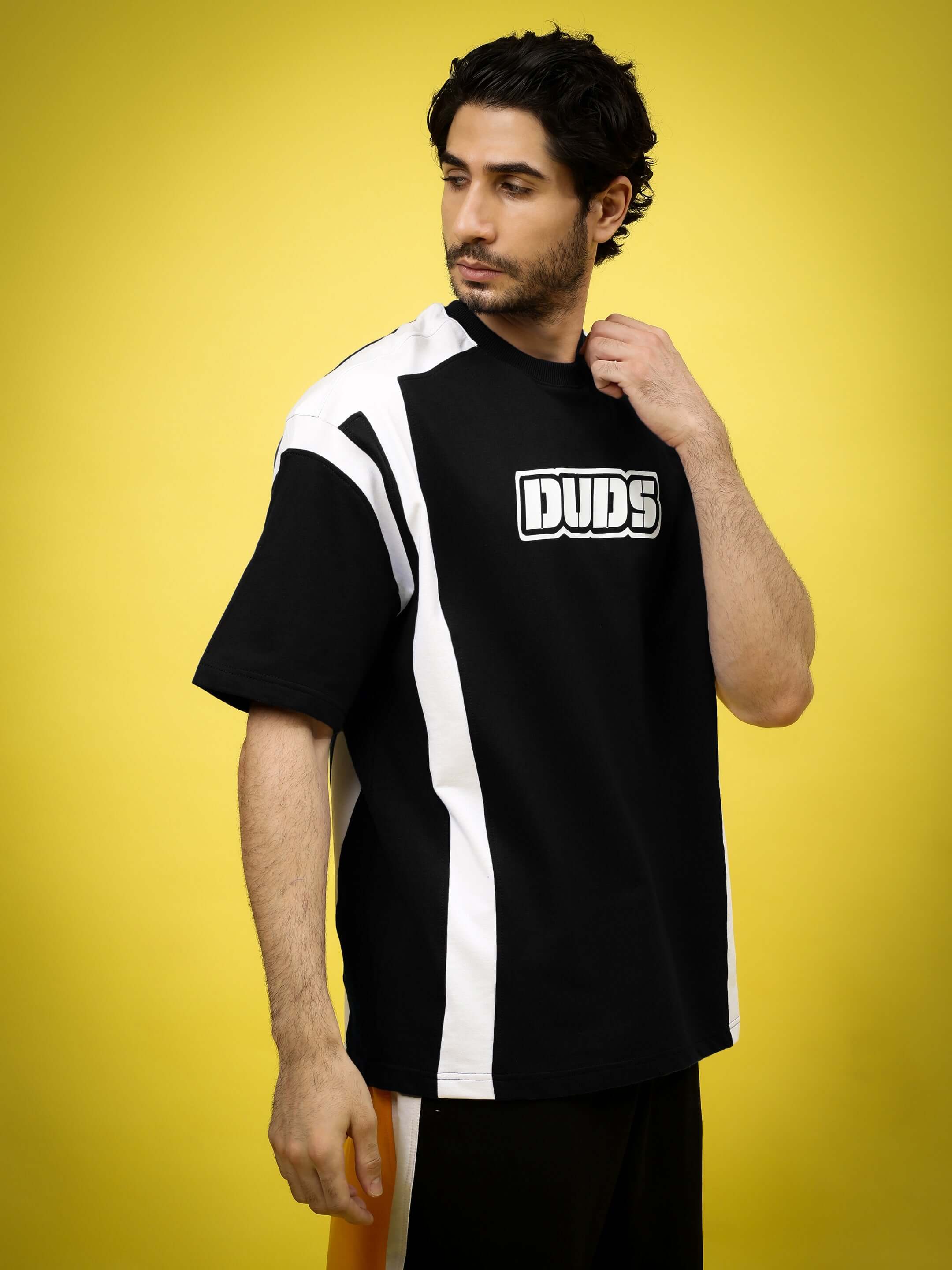 Man wearing a black and white oversized t-shirt with 'Duds' logo, standing against a yellow background.
