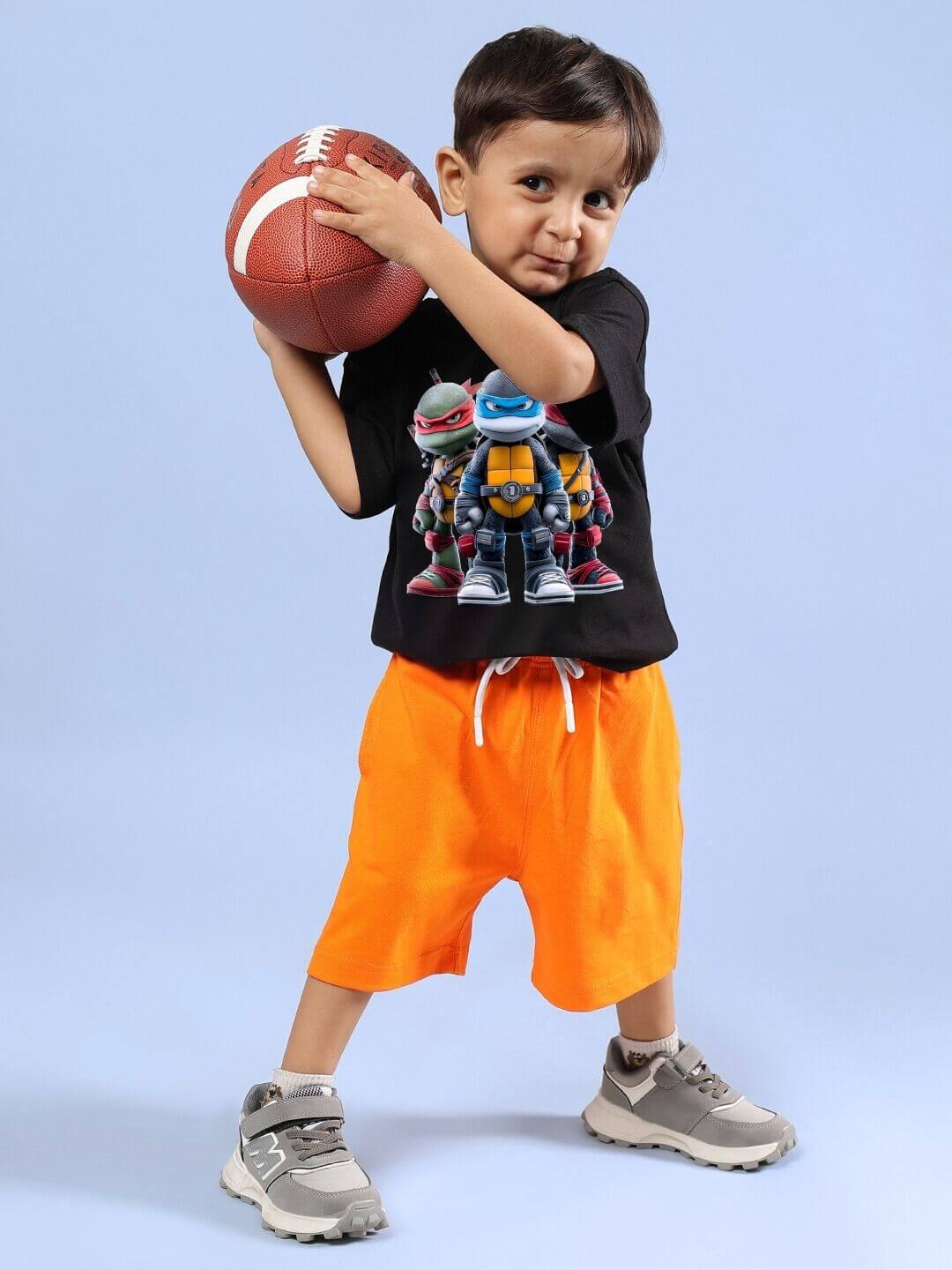 Child wearing Ninja Turtles black T-shirt and orange shorts holding a football, displaying action-themed style for boys and girls.