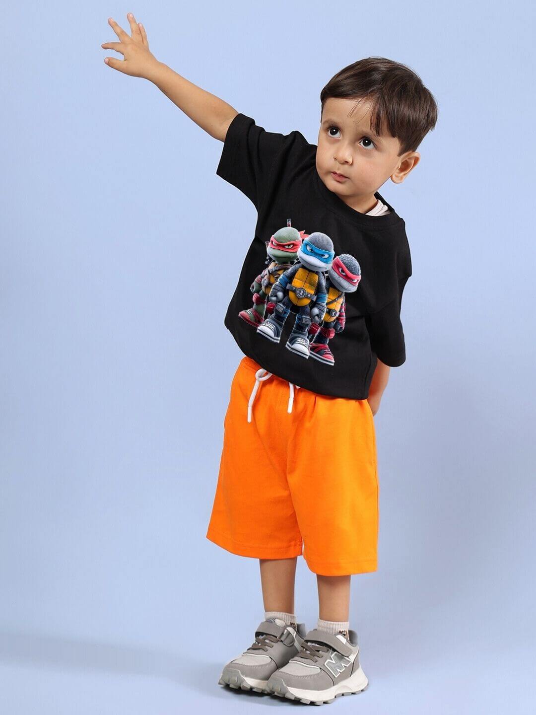 Child wearing Ninja Turtles co-ord set with black T-shirt and orange shorts, standing on a blue background.