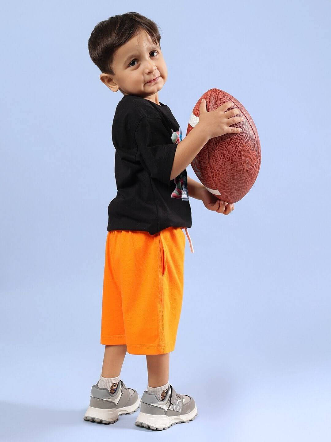 Child wearing Ninja Turtles black T-shirt and orange shorts, holding a football, showcasing boys and girls co-ord set.