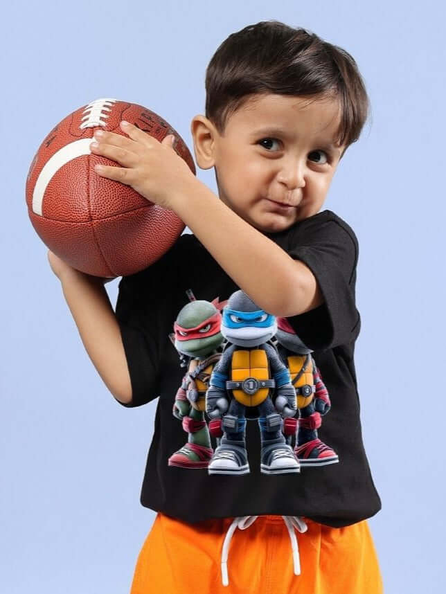 Child wearing NINJA TURTLES T-SHIRT FOR BOYS AND GIRLS BLACK holding a football.