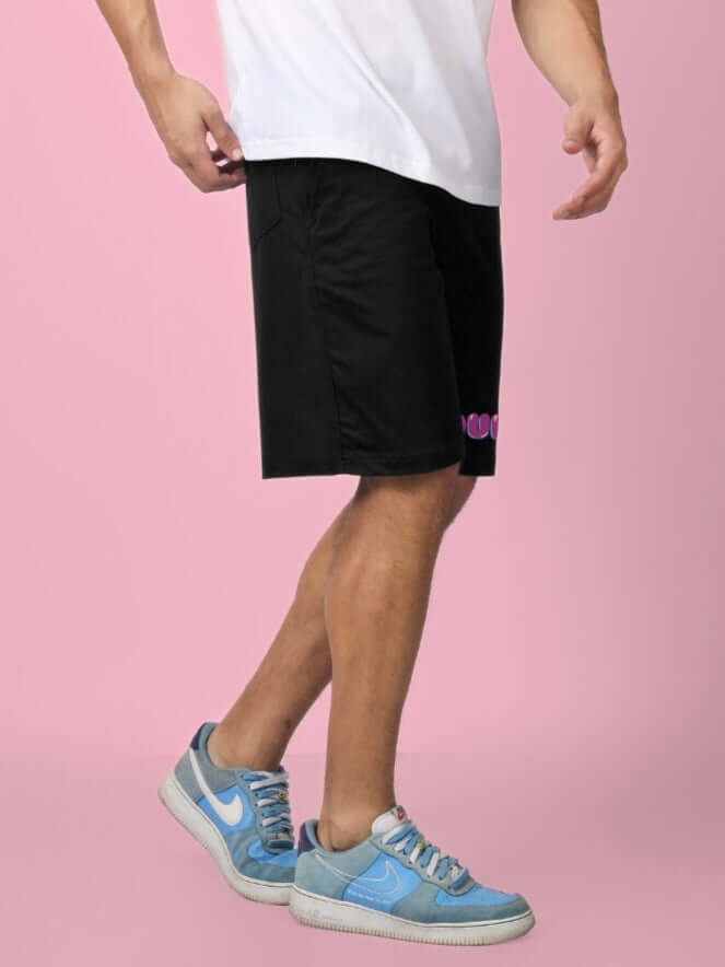 Person wearing NO POS SUMMER SHORTS COTTON BLACK with blue sneakers against a pink background.