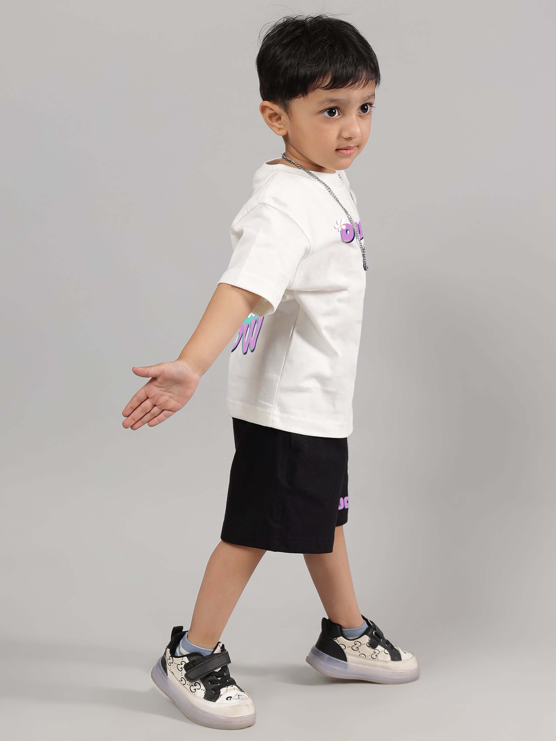 A young child wearing an off-white NO POS WOW T-shirt from DUDS, paired with black shorts and casual sneakers.