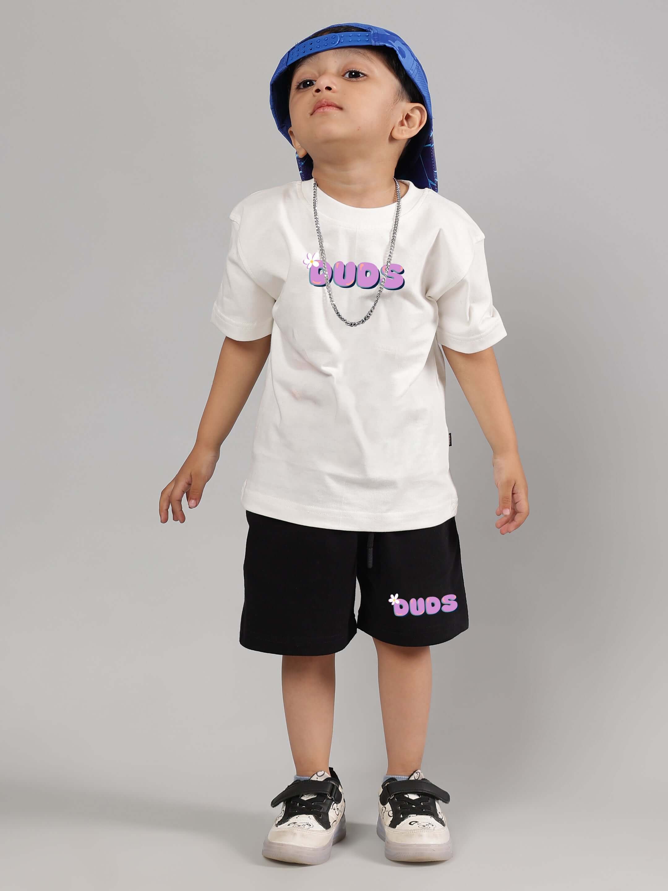 Child wearing NO POS WOW T-shirt in off-white by DUDS, customized fit available, paired with black shorts and blue hat.