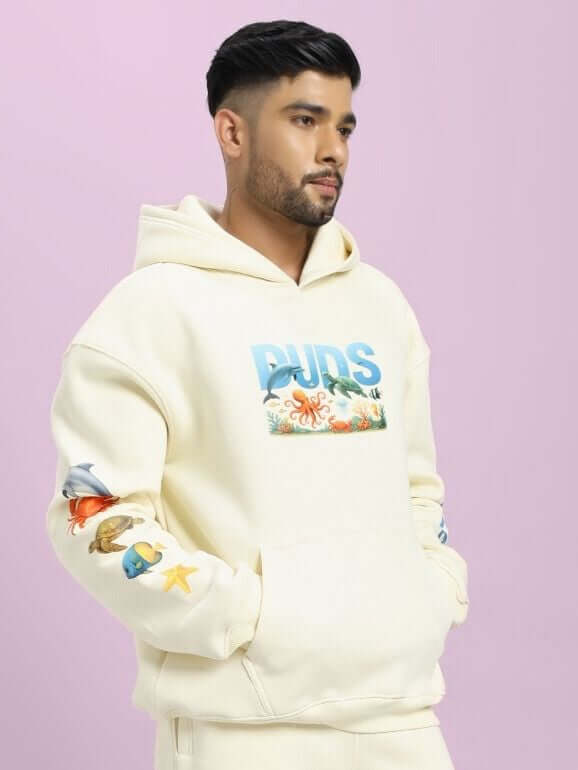 Man wearing OCEAN BUDDIES OVERSIZED FLEECE HOODIE OFF-WHITE with ocean-themed graphics on sleeves and front.