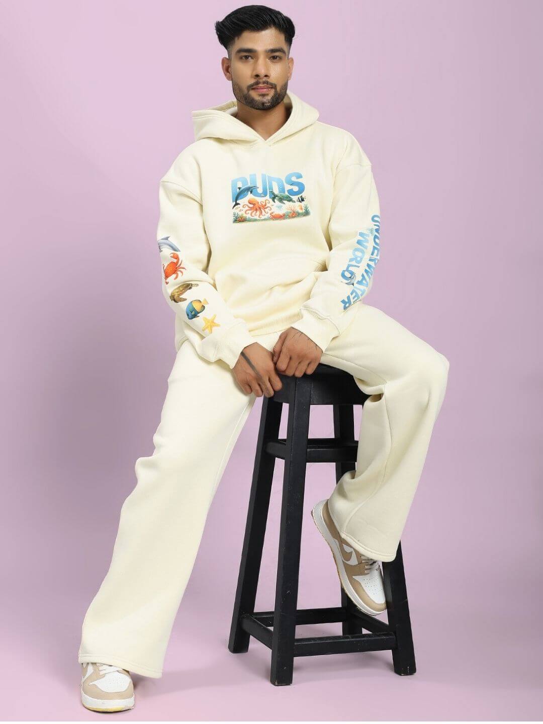Model wearing OCEAN BUDDIES JOGGER OFF-WHITE with sea life graphics, sitting on a stool, showcasing casual comfort and style.