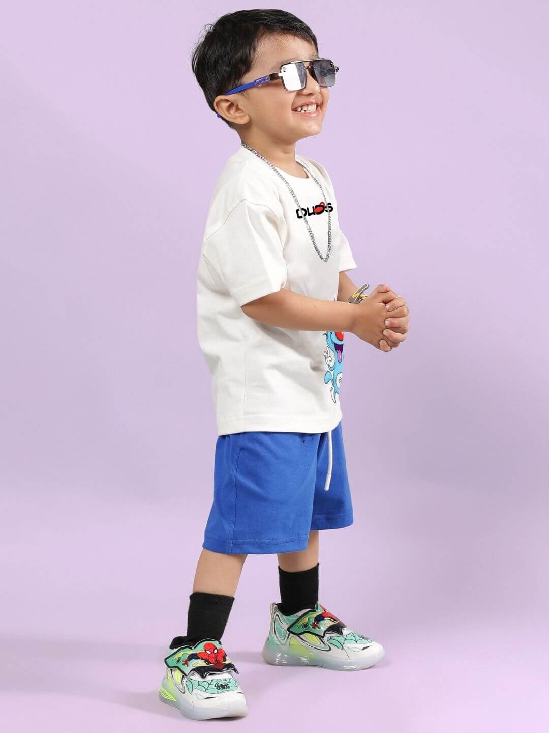 Child wearing Oggy printed co-ord set with off-white T-shirt and blue shorts, accessorized with sunglasses and sneakers, on a light purple background.