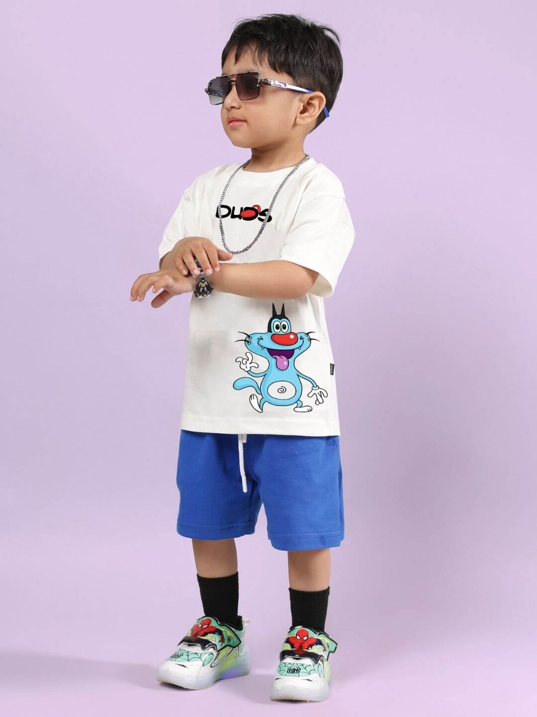 Boy wearing Oggy printed off-white T-shirt with blue shorts from co-ord set, stylish and comfy for kids, against a purple background.