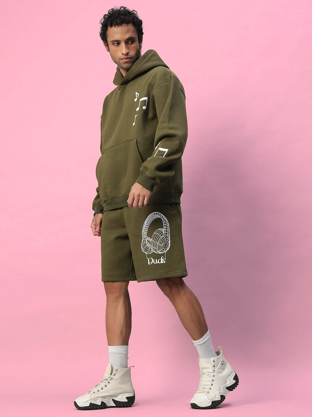 Model wearing OPERA FLEECE CO-ORD OLIVE GREEN set, featuring hoodie and shorts, with white shoes, on a pink background, highlighting casual style.