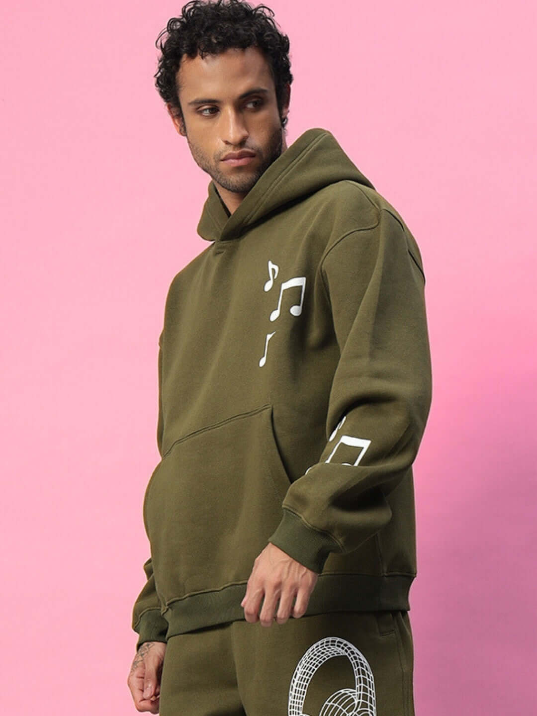 Model wearing OPERA FLEECE HOODIE OLIVE GREEN with musical notes design against pink background.