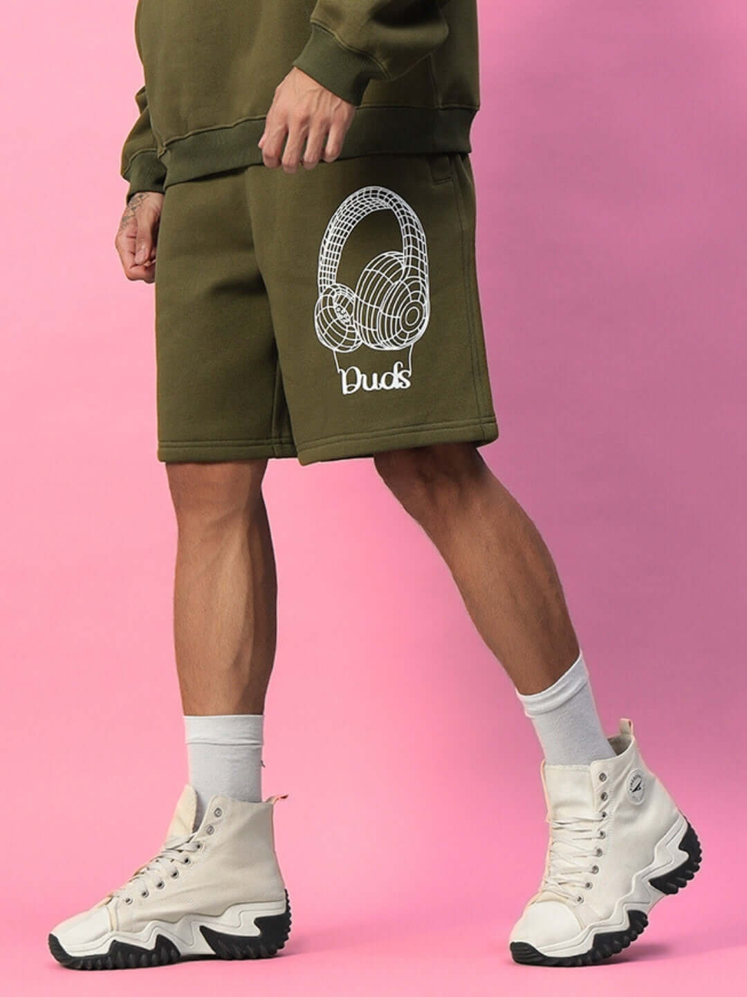 Person wearing OPERA FLEECE SHORTS OLIVE GREEN with headphone design, paired with white sneakers against pink background.