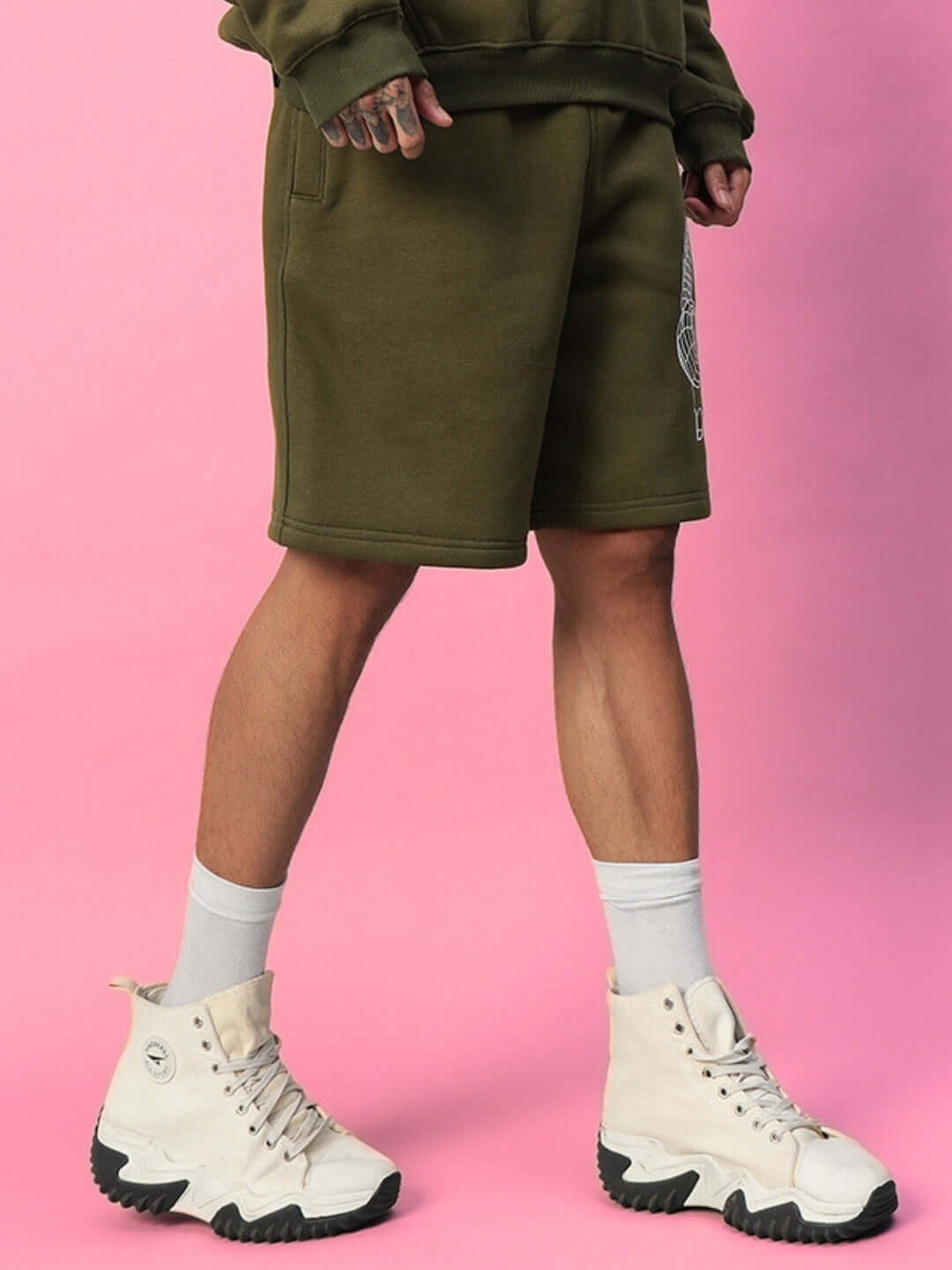 OPERA FLEECE SHORTS OLIVE GREEN worn with casual sneakers against a pink background.