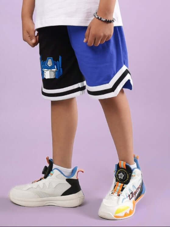 Child wearing Optimus Prime black-blue shorts from DUDS with white sneakers, showing front view on a purple background.