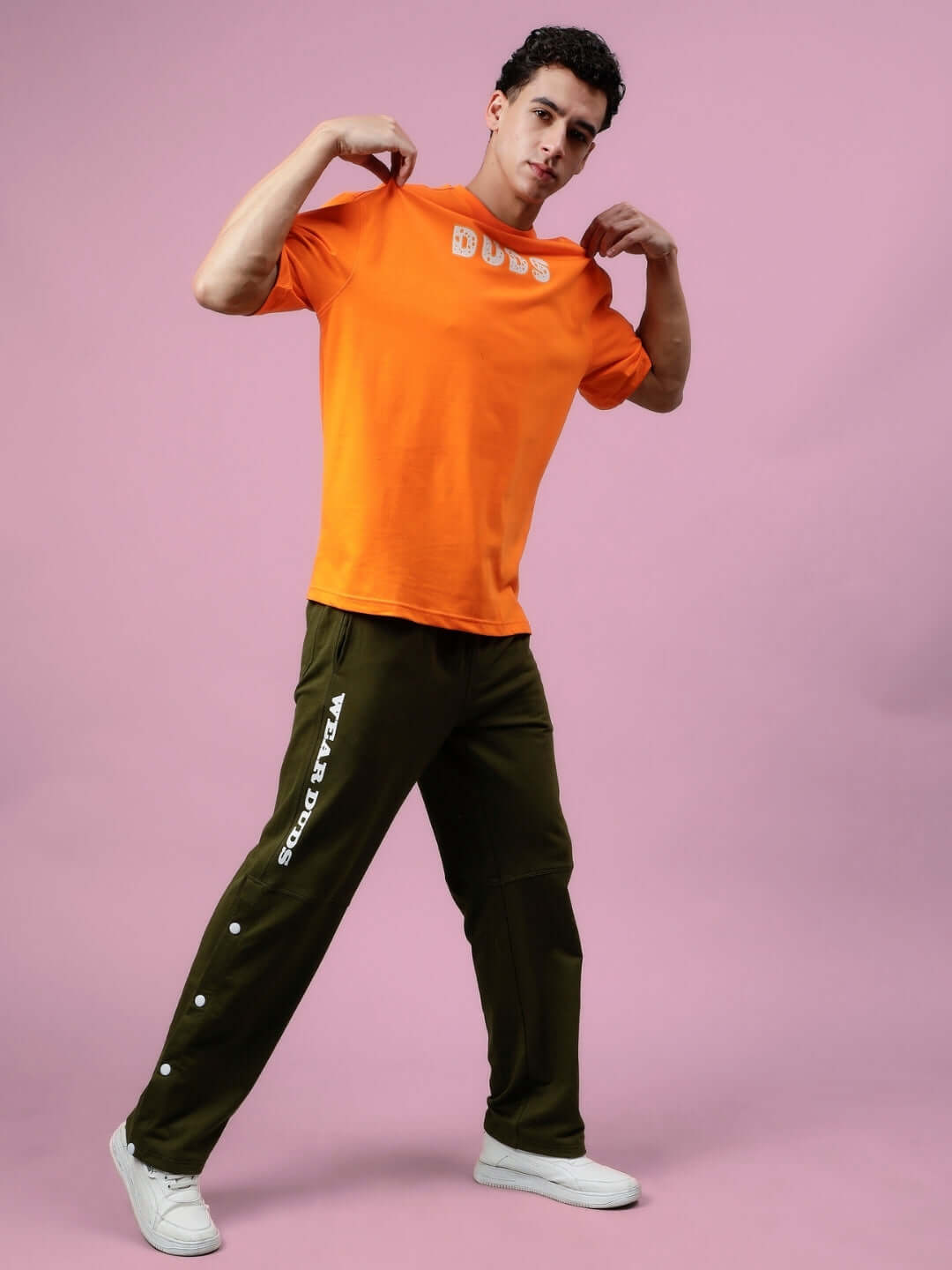 Model wearing COURAGE oversized orange t-shirt by DUDS with custom fit option, paired with olive pants on a pink background.