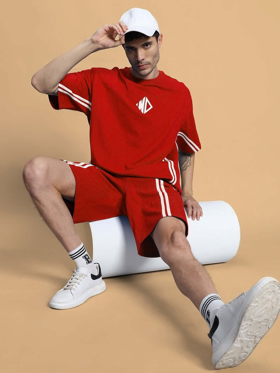 Model wearing OUTLOOK REGULAR FIT SHORTS RED with matching top and white sneakers, sitting on a white cylinder against a beige backdrop.