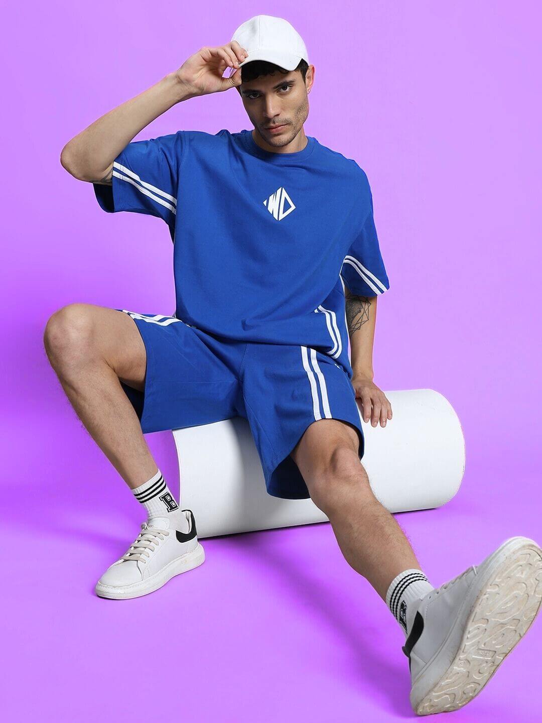 Model wearing OUTLOOK SUMMER CO-ORD SHORTS SET in royal blue with white sneakers against a purple backdrop.