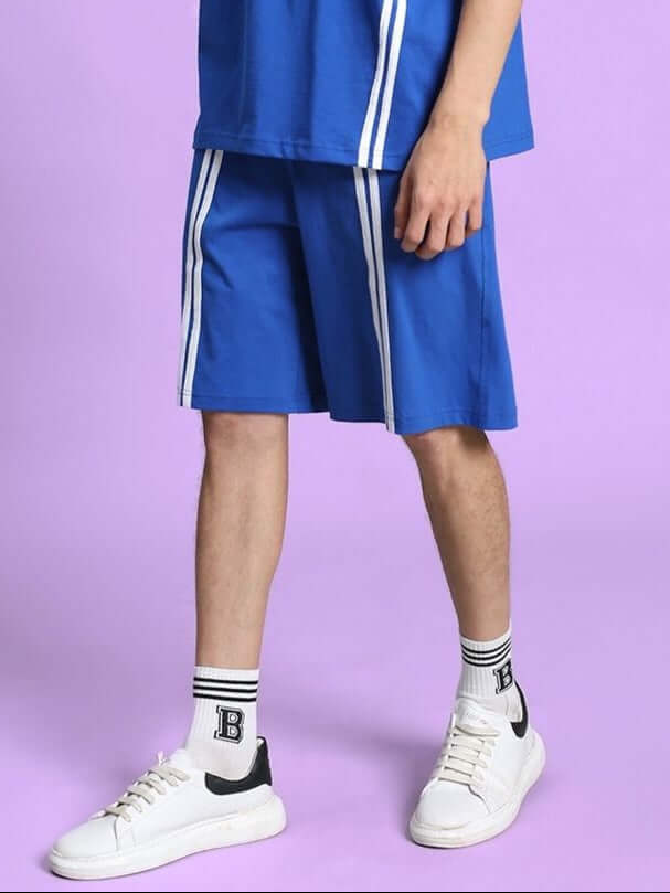 Man wearing OUTLOOK REGULAR FIT SHORTS ROYAL BLUE with white stripes, paired with white sneakers against a purple background.