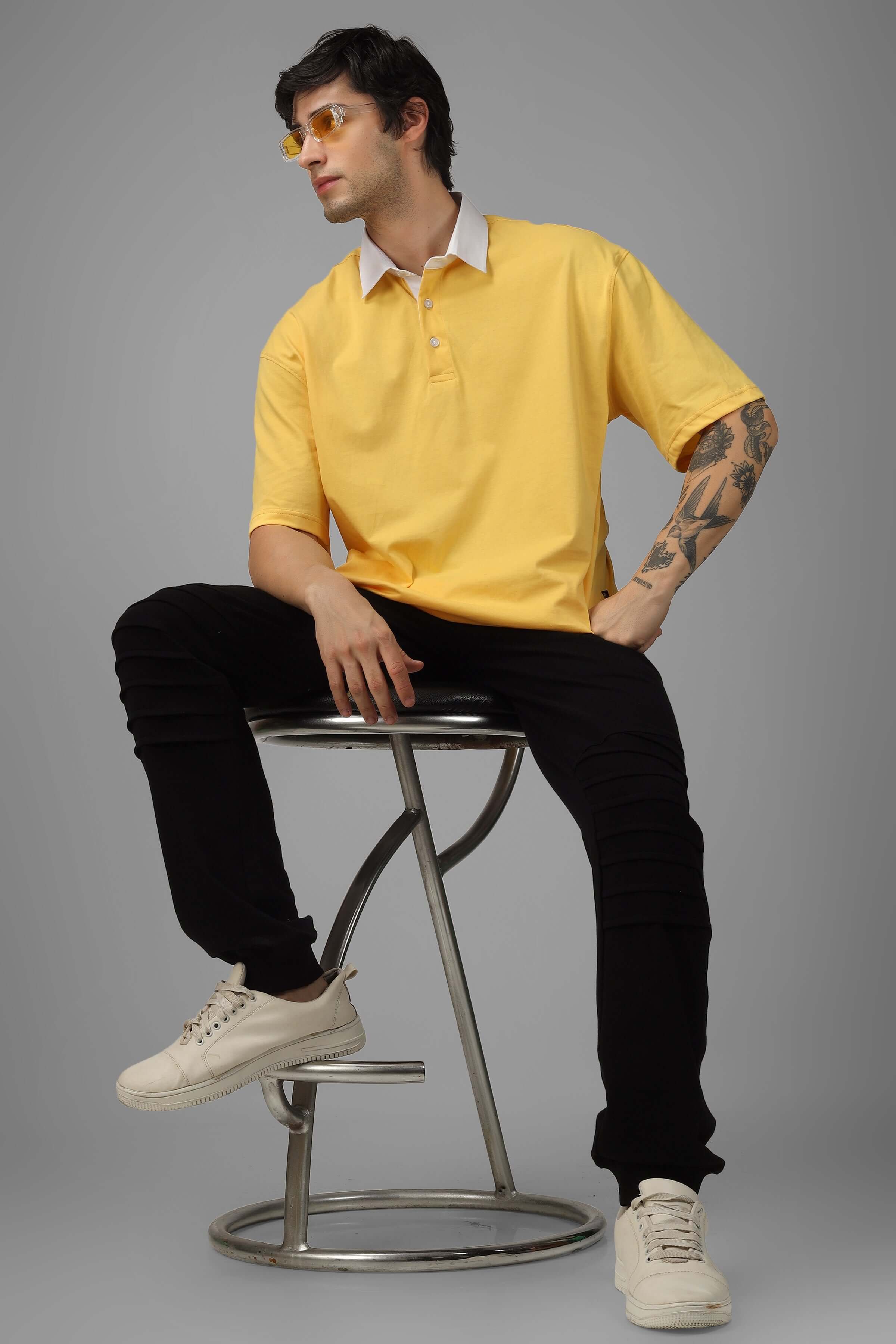Model wearing SAGOL POLO oversized yellow polo t-shirt with half sleeves, sitting on a stool; premium 100% cotton fabric.