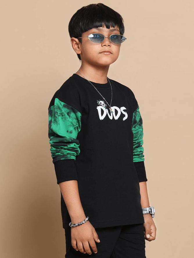 Child wearing a black oversized sweatshirt with 'DUDS' logo, featuring green patterned sleeves and sunglasses, showcasing trendy style.