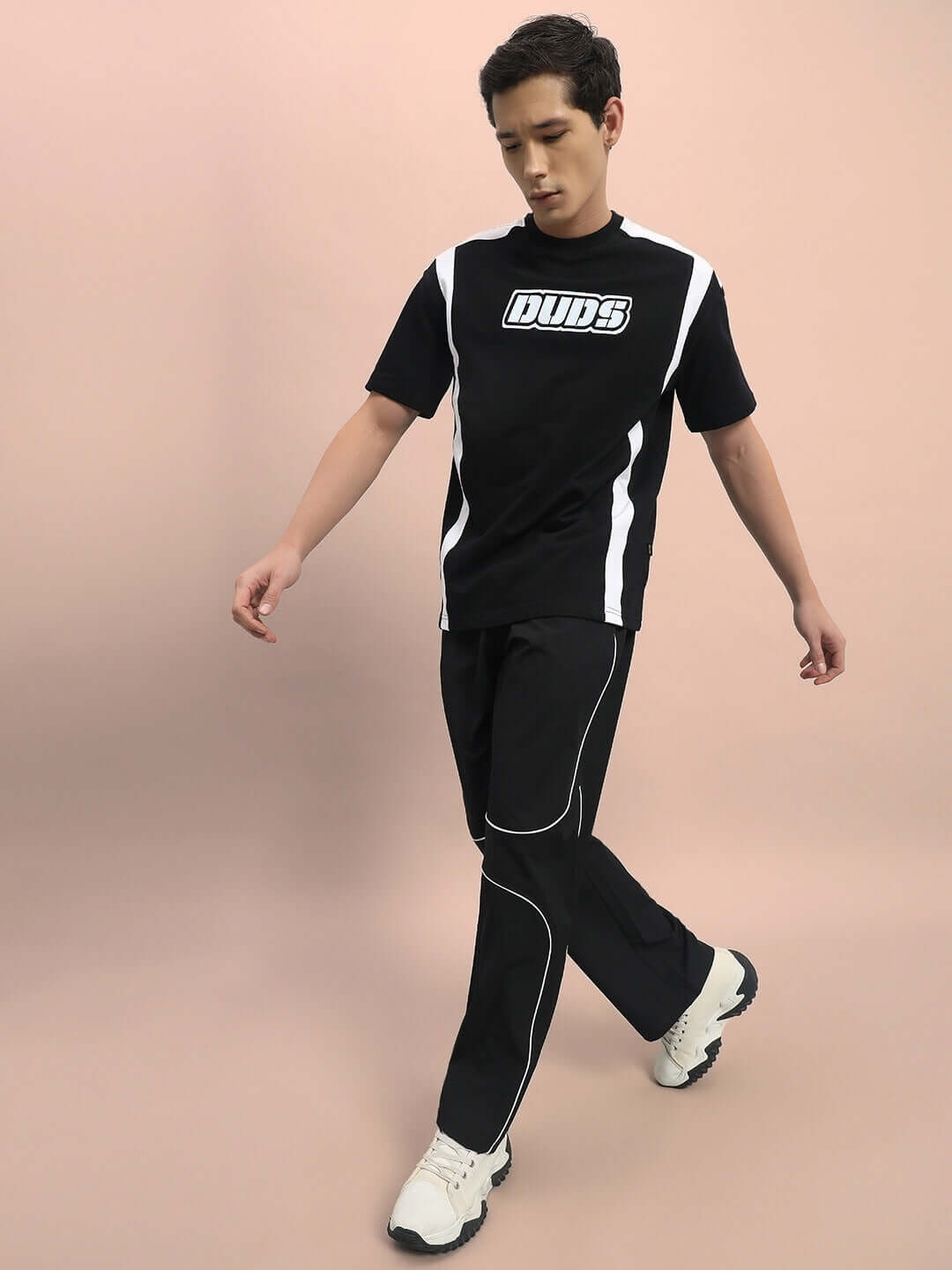 Model wearing DUDS black parabolic parachute cargo pants with white piping, paired with a black and white logo t-shirt, and white sneakers.