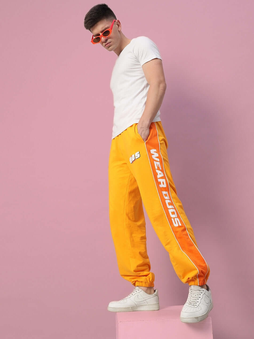 Man wearing yellow-orange PEPPY JOGGERS by DUDS, standing on a pink platform, sunglasses complementing the sporty ensemble.