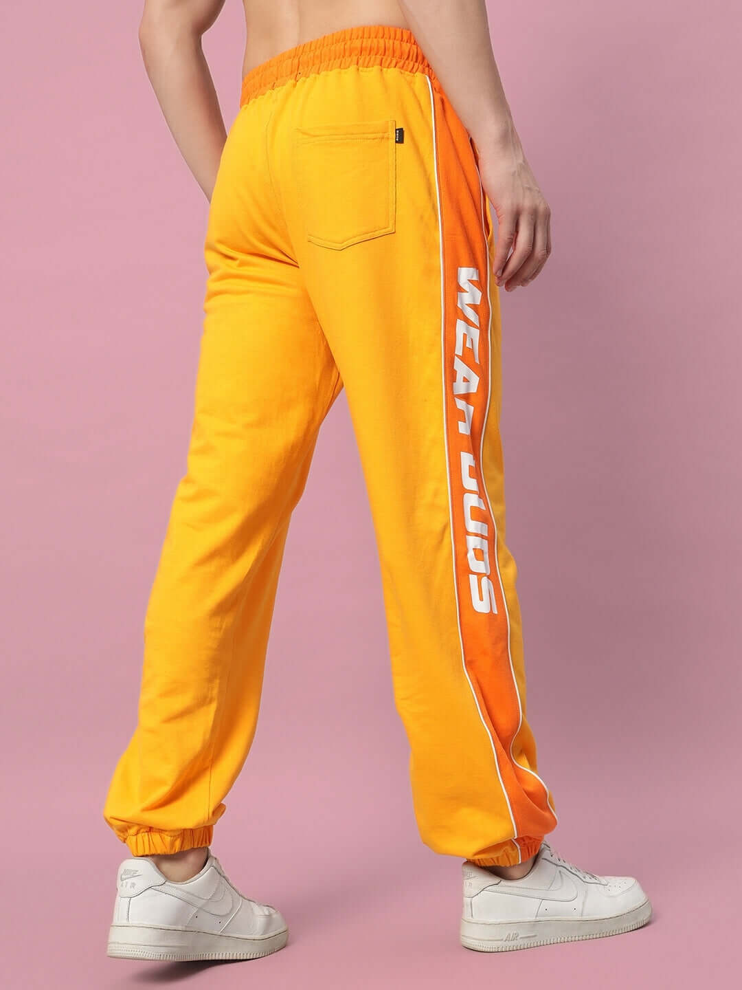 Yellow-orange Peppy Joggers with 'WEAR DUDS' text on side, shown from the back, paired with white sneakers, on a pink background.