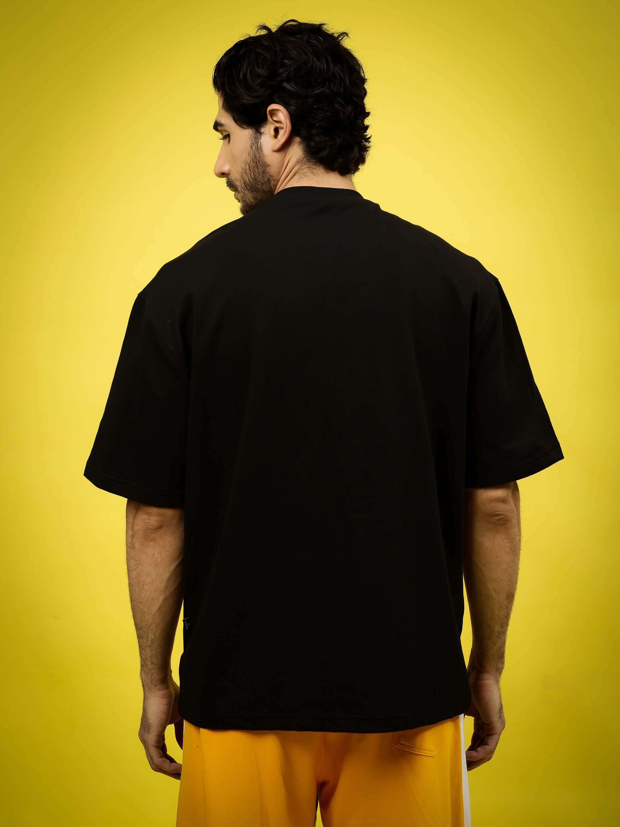 Back view of PERILOUS OVER-SIZED T-SHIRT BLACK on model, showcasing relaxed fit and bold design against a yellow background.