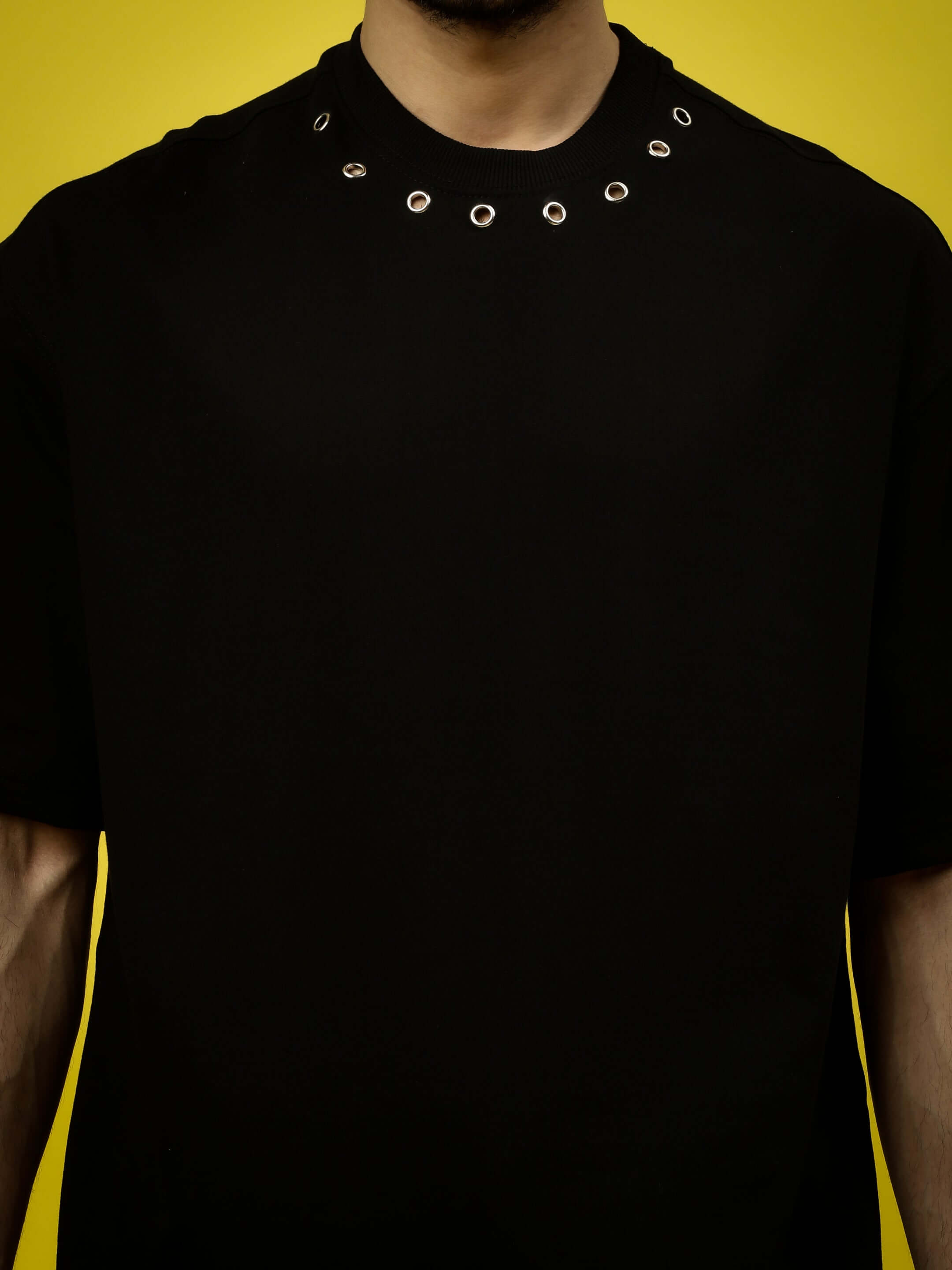 Man wearing PERILOUS OVER-SIZED T-SHIRT BLACK with round neck and metal ring details against yellow background.