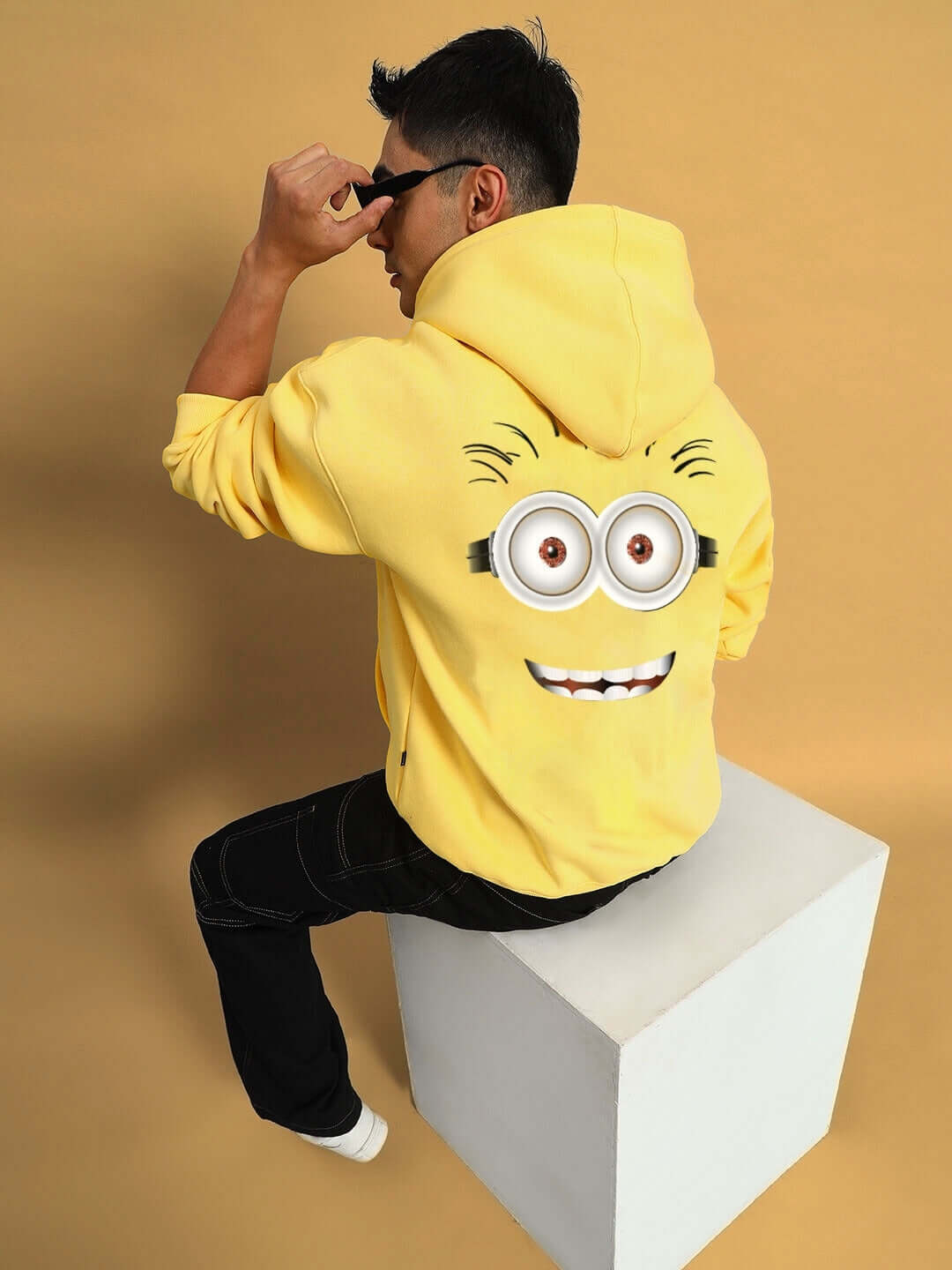 Man wearing yellow oversized hoodie with Minion design, sitting on a block, promotional image for customizable PHIL MINION hoodie.