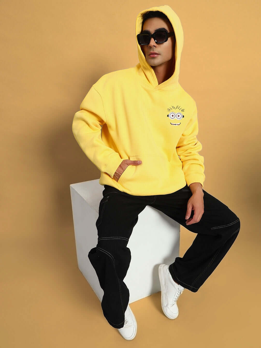 Model wearing PHIL MINION OVERSIZED FLEECE HOODIE YELLOW with black pants and white sneakers, sitting on a white block against a yellow background.