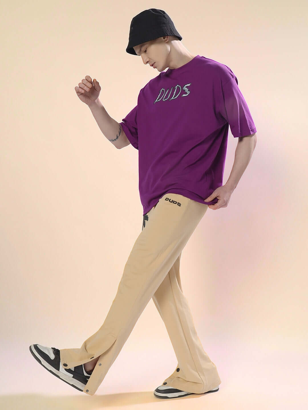Model wearing PHOENIX Digital Printed Joggers in beige, paired with a purple DUDS shirt and black hat.