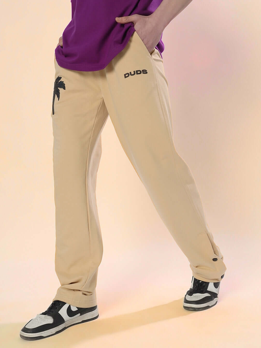 Man wearing beige digital printed joggers with palm tree design and black sneakers, hands in pockets.