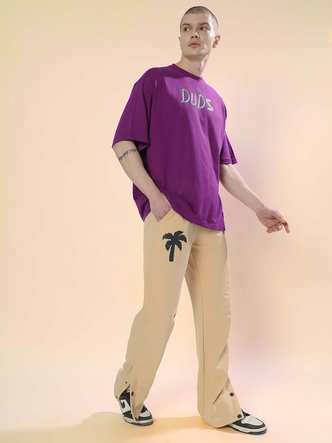 Model wearing Phoenix digital printed beige joggers with palm tree design, paired with a purple DUDS t-shirt.