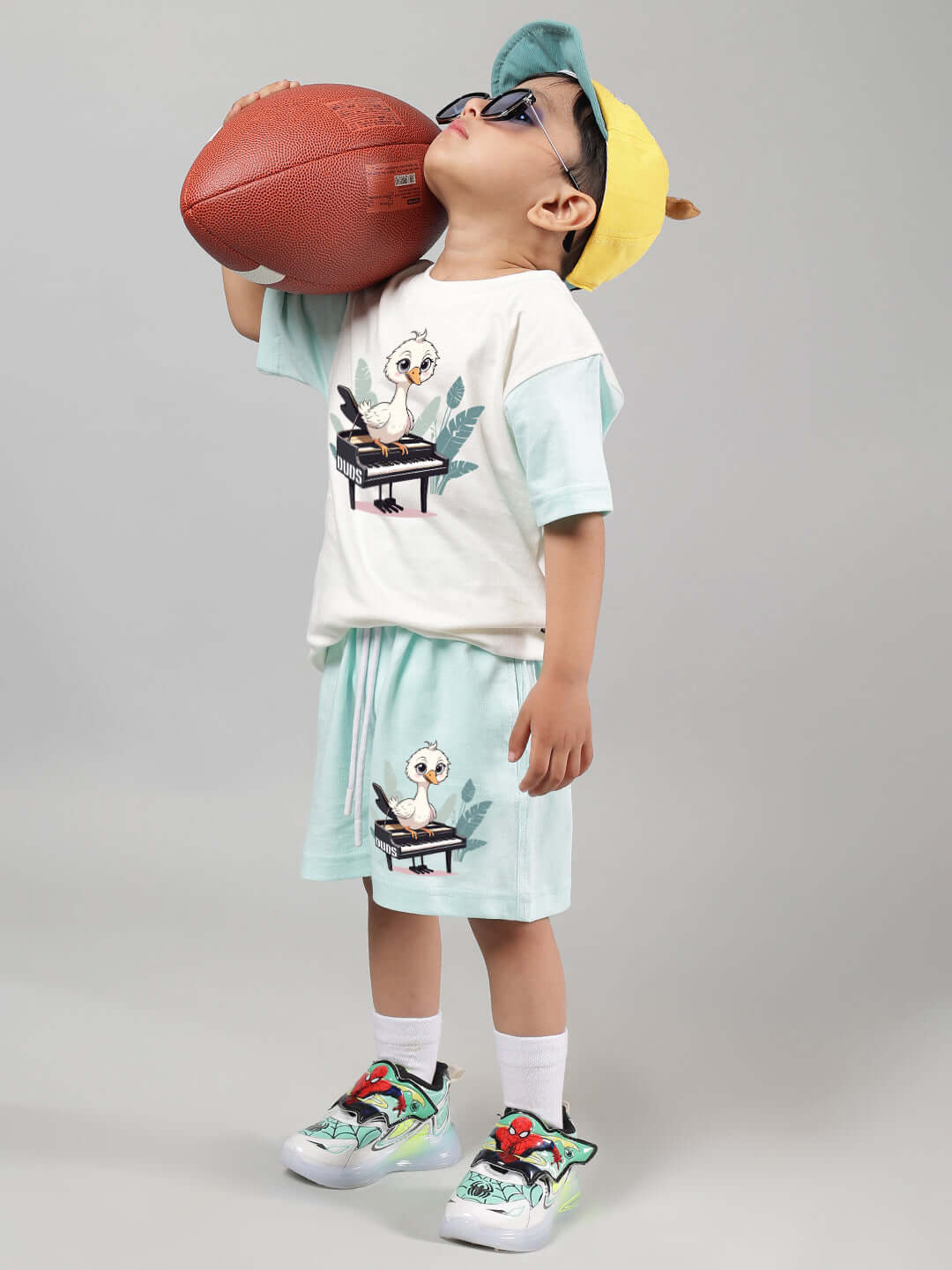 Child wearing an off white-blue Piano Bird T-shirt and shorts, holding a football, showcasing cute and playful style for boys and girls.