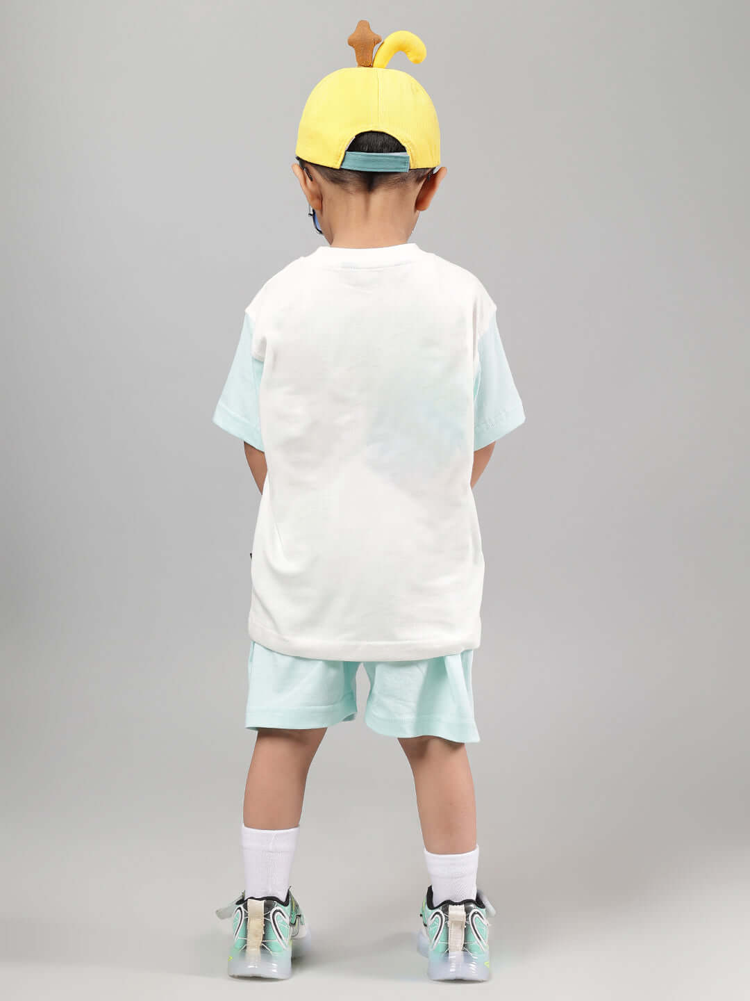 Back view of a child wearing an off-white and blue Piano Bird T-shirt, paired with a yellow cap, showcasing a casual fashion style.