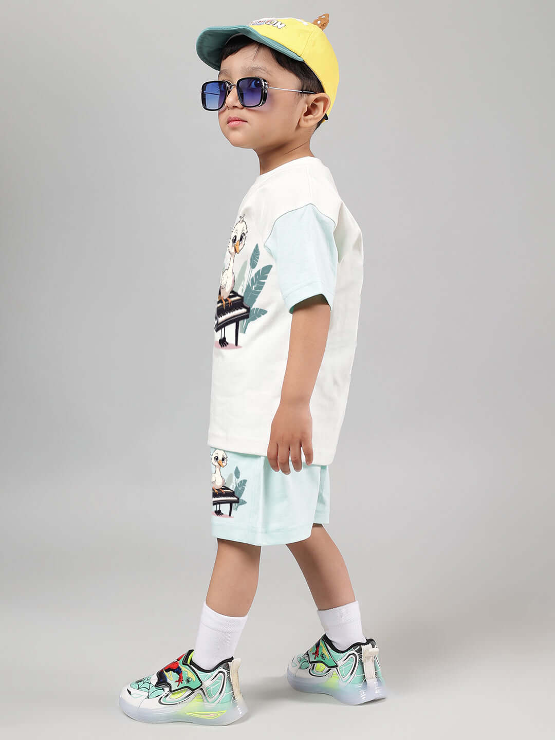 Child wearing off-white and blue piano bird t-shirt and shorts, styled with a yellow cap and sunglasses.