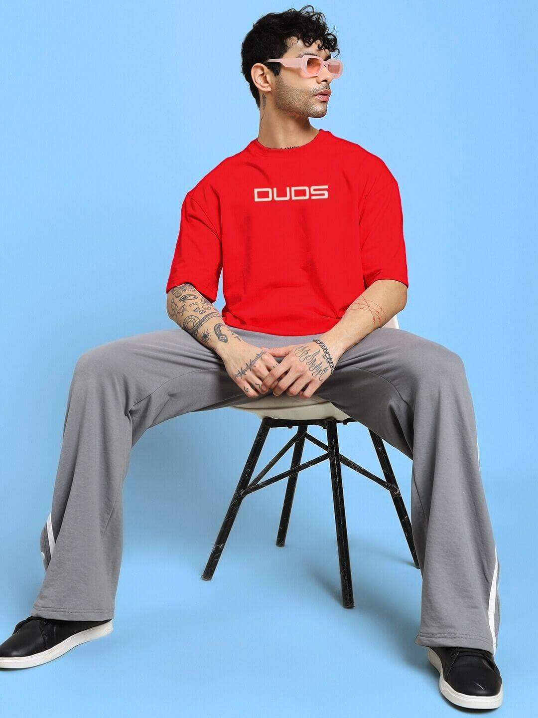 Man wearing Porsche Printed Over-Sized T-Shirt Red with grey pants, seated on a stool, in a casual pose against a blue background.