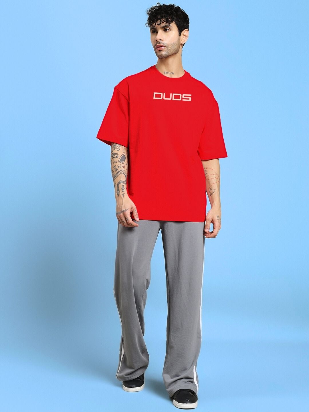 Man wearing a red PORSCHE PRINTED OVER-SIZED T-SHIRT RED with gray pants, standing on a blue background.