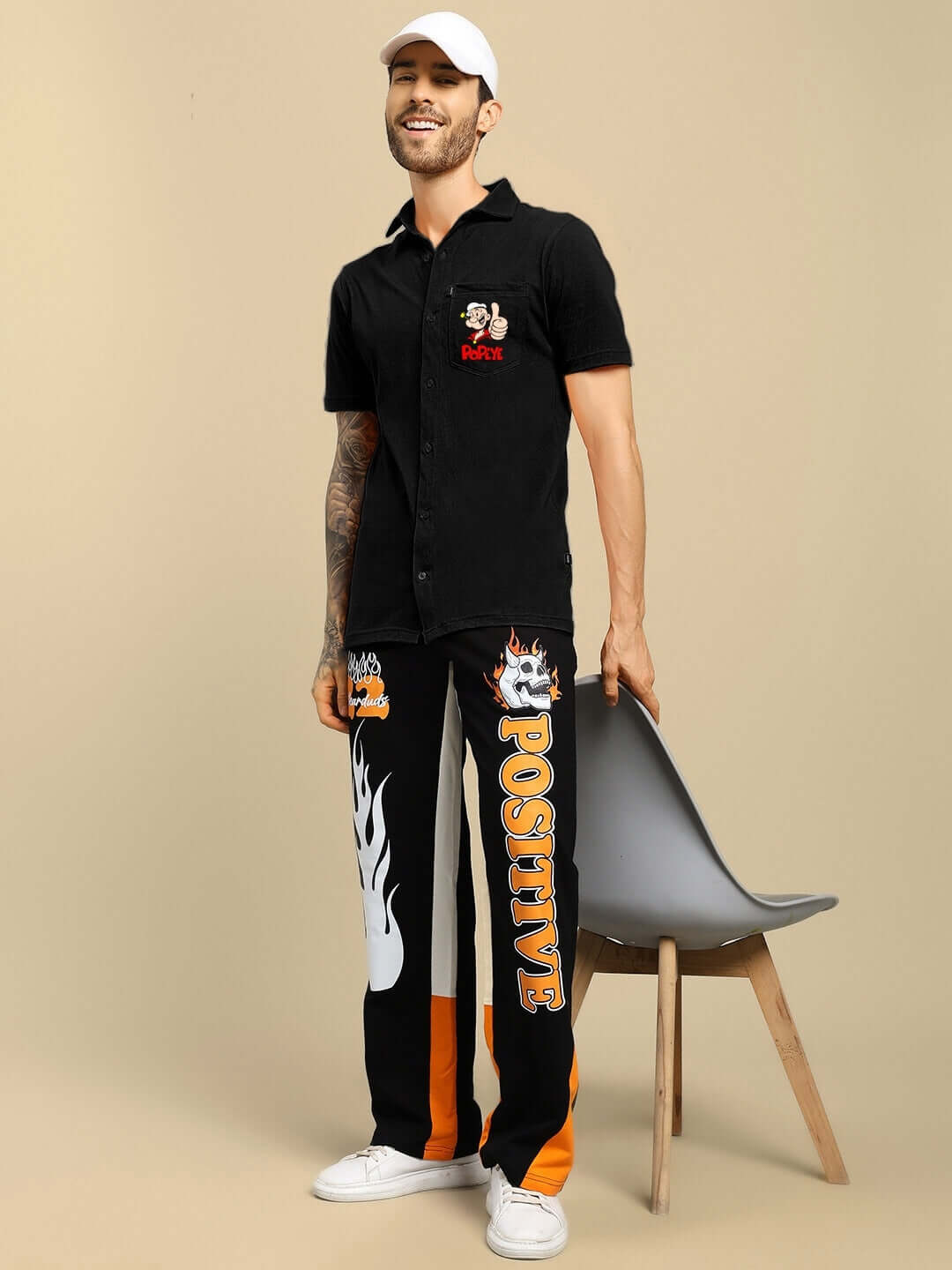 Man wearing black Popeye printed shirt and bold graphic pants, standing by a chair.