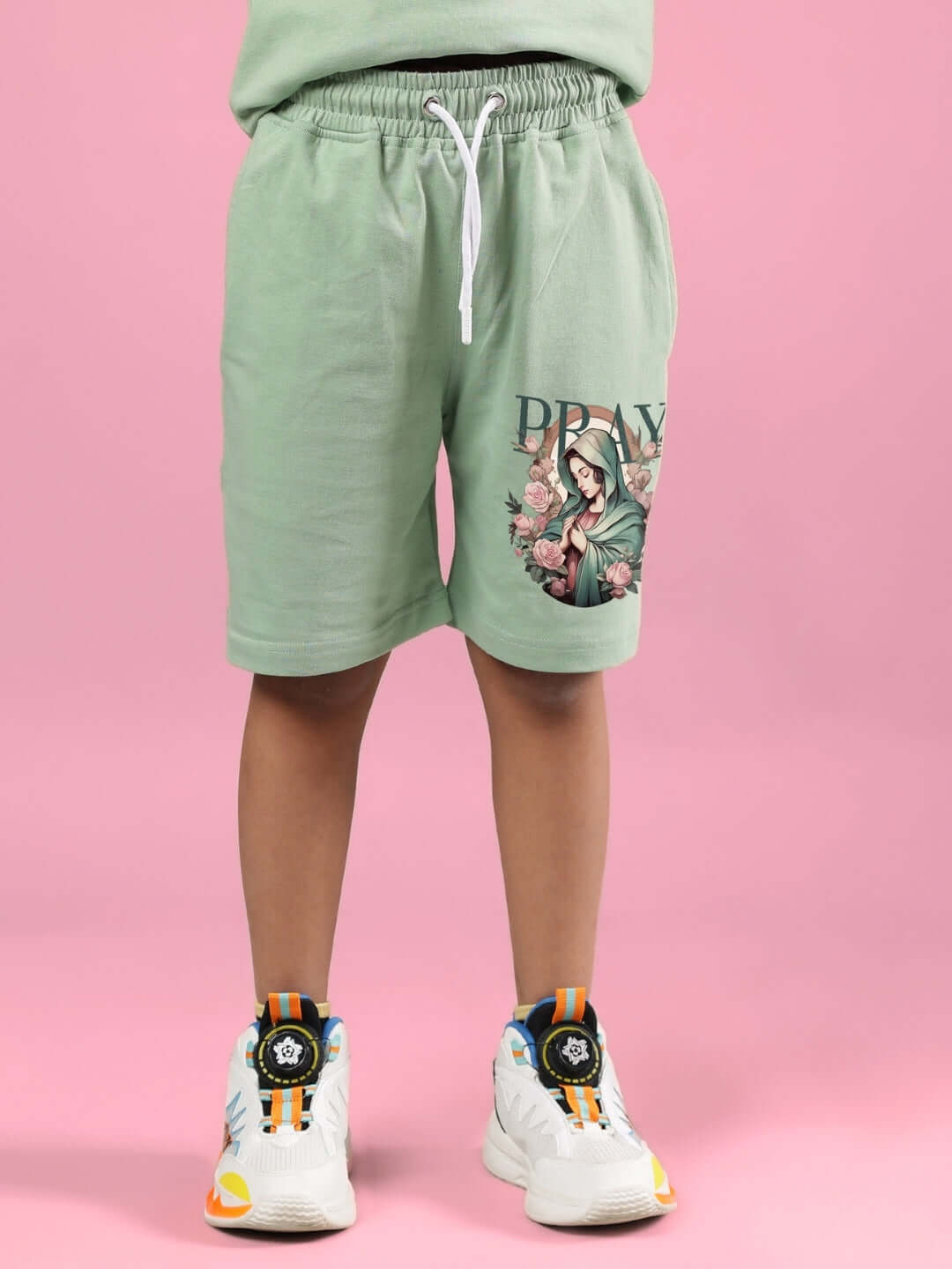 Child wearing PRAY & PEACE CO-ORD FOR BOYS & GIRLS NYANZA GREEN shorts, featuring a graphic design, on a pink background.