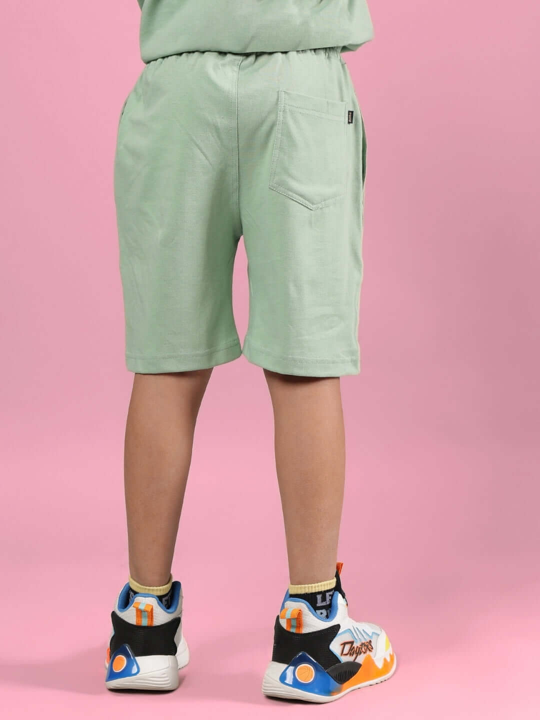 Rear view of child wearing PRAY & PEACE SHORTS FOR BOYS & GIRLS NYANZA GREEN standing against pink background.