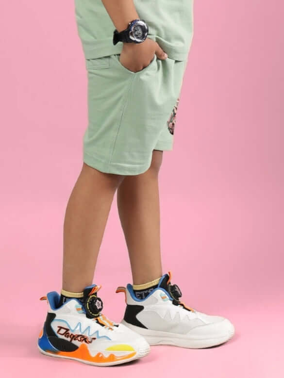 Boy in colorful sneakers wearing PRAY & PEACE SHORTS FOR BOYS & GIRLS NYANZA GREEN against pink background.