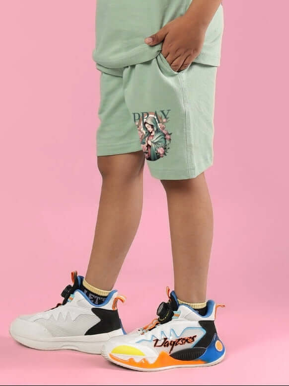 Child wearing PRAY & PEACE SHORTS FOR BOYS & GIRLS NYANZA GREEN with colorful sneakers on pink background.